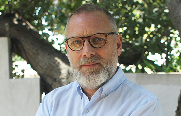 A man with short grey hair, a grey beard, and glasses wearing a light blue button-down shirt.