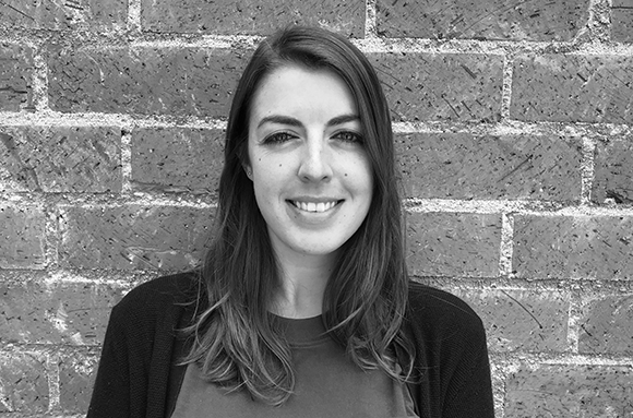 A smiling woman with long hair poses in front of a brick wall in a black and white portrait.