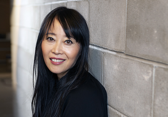 A woman with long black hair and bangs smiles while leaning against a light gray stone block wall.