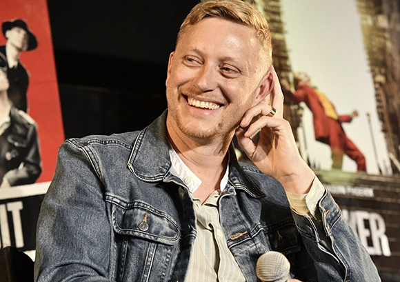 A smiling man with short blonde hair wearing a denim jacket and striped shirt.