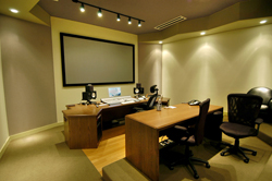 A media editing room featuring a large projection screen, wooden desks, computer monitors, and office chairs.