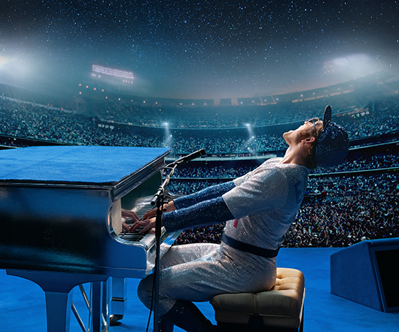 A person in a sparkling outfit plays a blue grand piano in a massive, crowded stadium under a starry sky.