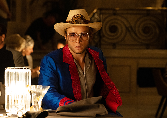 Man wearing a tan cowboy hat, large glasses, and a blue blazer with red trim sits at a dimly lit table.