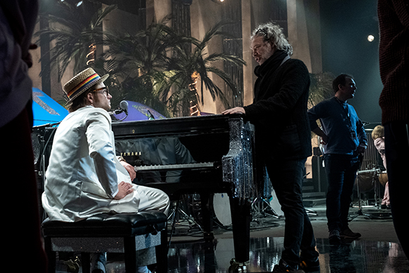 A man in a white suit and straw hat sits at a black grand piano while a man in black stands next to him.