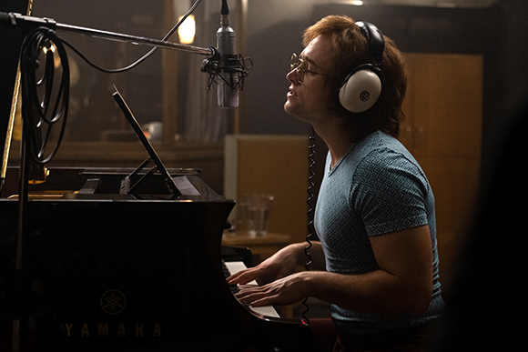 A man with glasses and headphones playing a black Yamaha piano in a dimly lit studio.