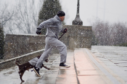 A man in grey athletic wear and a dark dog run down stone steps during a snowfall.