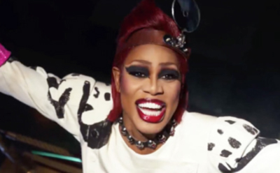 A person with bright red hair, heavy dark eye makeup, and red lipstick smiling broadly in a spotted outfit.