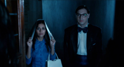 A woman in a blue shirt holds up a white veil while a man in a tuxedo and glasses stands beside her.