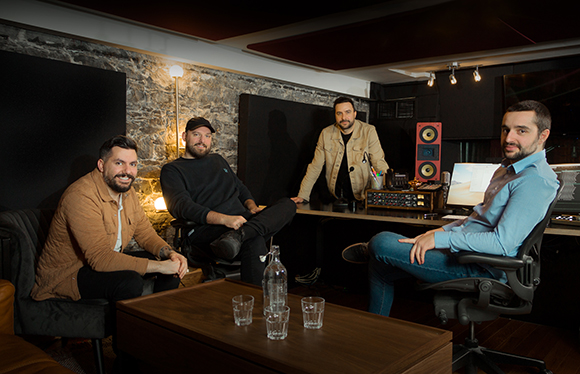 Four men sitting and standing in a professional music recording studio with equipment.