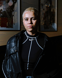 A person with short blonde hair looks forward, wearing a black leather jacket and a black Nike turtleneck.
