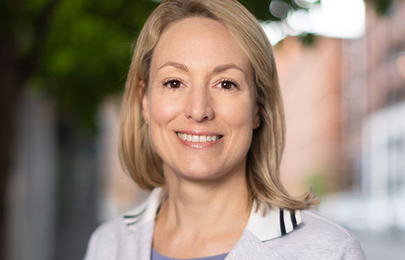 A smiling woman with blonde hair and light eyes wearing a white collared shirt.
