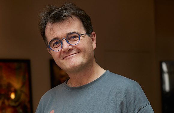 A man with short brown hair and blue-rimmed glasses smiling in a dimly lit room.