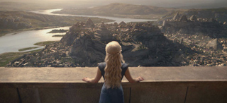 A woman with long blonde hair stands on a balcony overlooking a sprawling city and river valley.