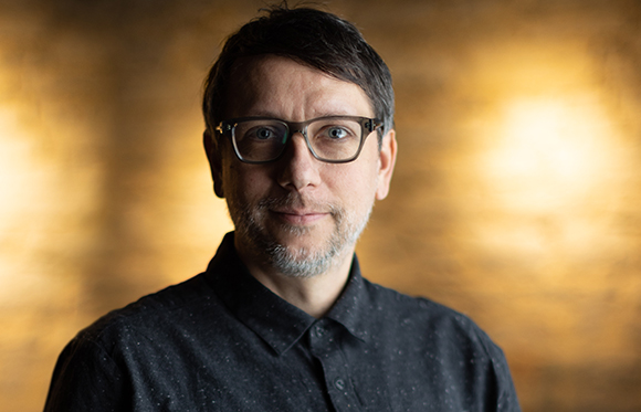 A man with short dark hair, glasses, and a salt-and-pepper beard wearing a dark button-down shirt.