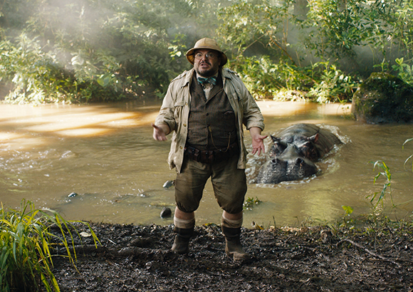 A man in a safari hat and tan jacket stands by a muddy river as a hippopotamus swims nearby.