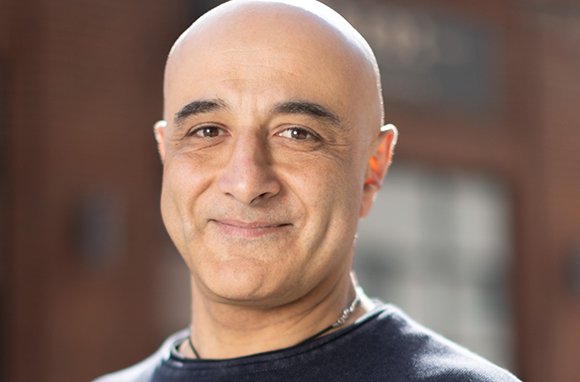 A close-up portrait of a bald man with dark eyes and a slight smile against a blurred background.