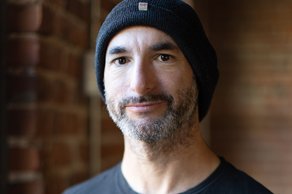 A man with a salt-and-pepper beard wearing a black beanie and black t-shirt against a brick background.