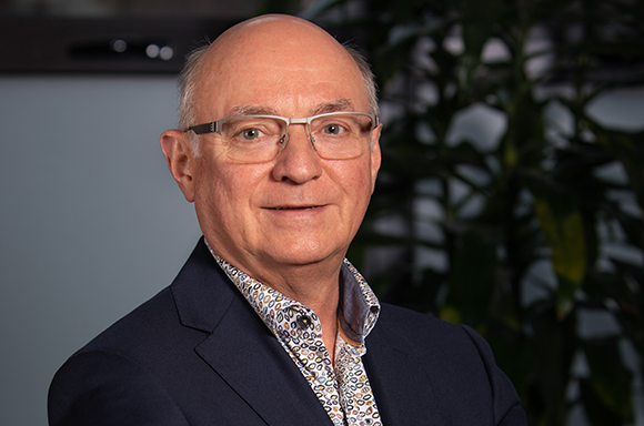 A bald man with glasses wearing a dark blazer and a patterned button-down shirt.