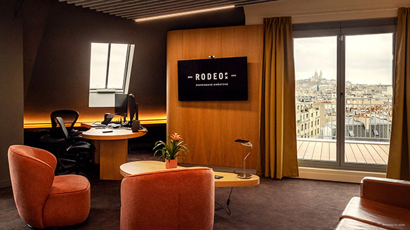 A modern office interior with a wooden desk, orange armchairs, and a large window overlooking a city skyline.