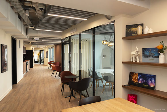 A modern office hallway with wood floors, glass-walled meeting rooms, and wooden shelves on the right wall.