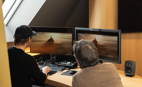 Two men sit at a wooden desk viewing a landscape image on two large computer monitors.