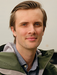 A man with light brown hair and blue eyes smiling slightly against a neutral background.