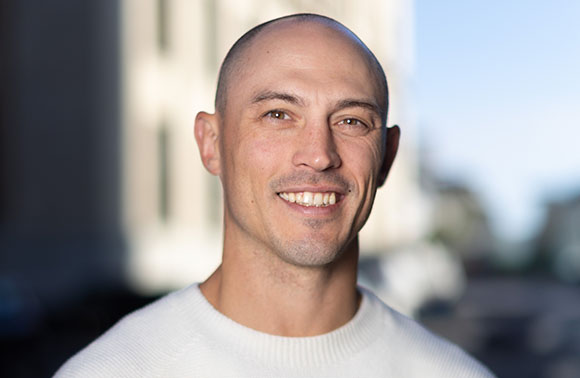 A bald man with a light complexion smiles at the camera while wearing a white textured sweater.