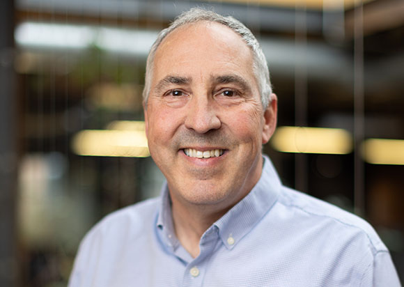 A smiling middle-aged man with short graying hair wearing a light blue button-down shirt.