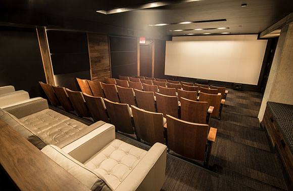 Rows of wooden and upholstered theater seats facing a large blank projection screen in a dark room.
