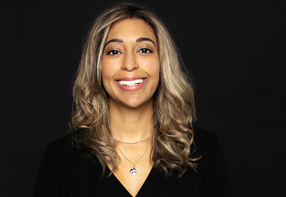 A smiling woman with wavy, highlighted hair wearing a black blazer against a black background.