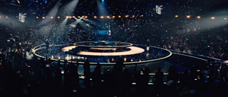 A circular, glowing stage in a dark arena surrounded by a large, silhouetted crowd under bright spotlights.