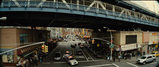 An elevated train track spans over a city street filled with cars, buses, and pedestrians.