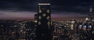 A dark cityscape at night with a tall skyscraper illuminated by scattered lights under a dim sky.