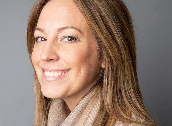 A smiling woman with long brown hair looking toward the camera against a gray background.