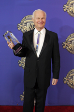 A man in a black tuxedo and white shirt holds a trophy against a blue background with gold logos.