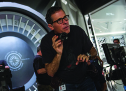A man wearing glasses and a black t-shirt holds a camera while looking off-camera on a film set.