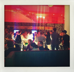 A crowd of people stand and mingle in a venue with red lighting and a staircase in the background.