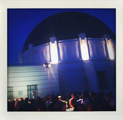 The Griffith Observatory building at dusk, illuminated by lights, with a crowd of people visible in front.