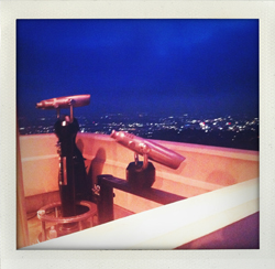 Two antique telescopes on a balcony with a city skyline in the background at twilight.