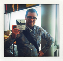 Man wearing glasses and a checkered shirt holds up a conference badge. He has a dark strap across his chest.
