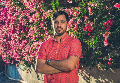 A man with dark hair and a beard stands with arms crossed in front of a wall of pink bougainvillea.