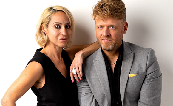 A blonde woman in a black sleeveless top leans her arm on a man wearing a grey blazer and black shirt.