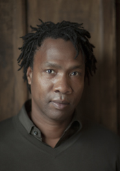 A man with dark dreadlocks looking directly at the camera against a wooden background.