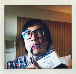 A man with glasses and a plaid shirt bites on a lanyard with a conference badge that reads Terence Lee.