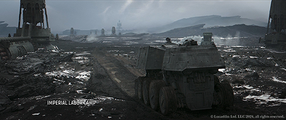A heavy industrial vehicle sits on a muddy, dark terrain under a cloudy sky near large metal structures.
