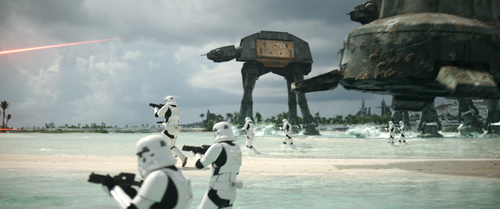 Stormtroopers advance across a sandy beach toward large, four-legged walkers under a cloudy sky.