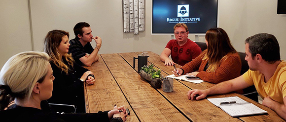 Four people sit around a long wooden table in a meeting room with a screen in the background.