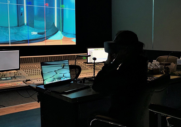 A person in silhouette wearing a VR headset sits at a desk with a laptop and mixing console in a dark studio.