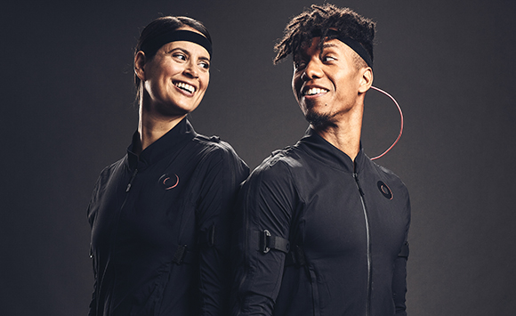 A woman and a man in black athletic wear and headbands smiling at one another against a dark background.