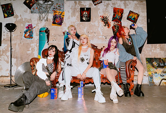 Five women in stylish streetwear pose together in a room decorated with colorful posters and graffiti.